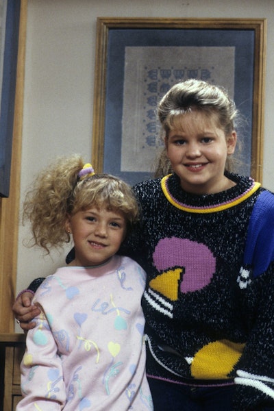 Young Jodie Sweetin and Candace Cameron Bure wearing scrunchies with ponytails on the set of 'Full House.'