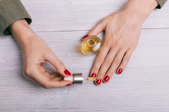 A person with red nail polish applies cuticle oil to their fingernails.