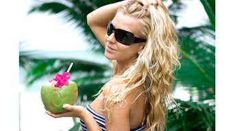 Woman in sunglasses holds coconut in hand.