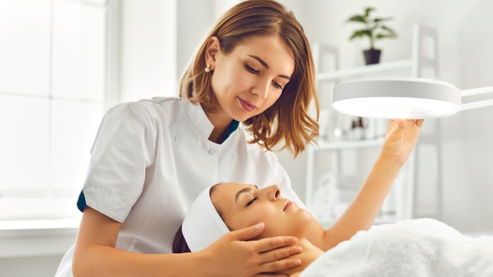 Cosmetologist directing lamp to woman for facial treatment