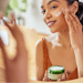 medium-toned woman applying natural skin cream in mirror smiling