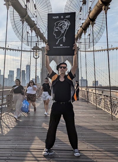 Damian Monzillo poses with Hair is Art campaign poster on the Brooklyn Bridge in New York.