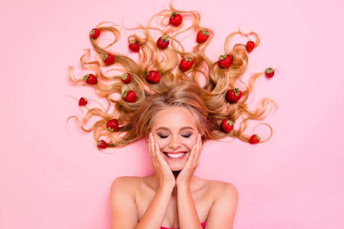 A woman with strawberry blonde hair lays with her hair splayed out with strawberries around it on a pink background. She is smiling, and her hands are on either of her cheeks.
