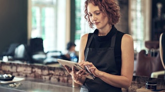 Woman with short, curly red hair in a hair salon reading a tablet.