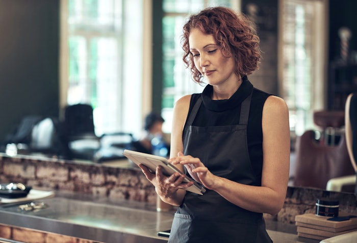 Woman with short, curly red hair in a hair salon reading a tablet.