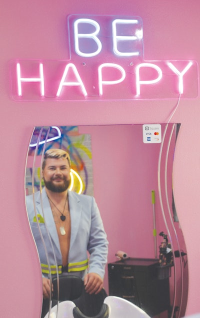 Willy Millard poses at salon chair. You can see his reflection in a mirror. Above the mirror is a neon sign that says 'BE HAPPY.'