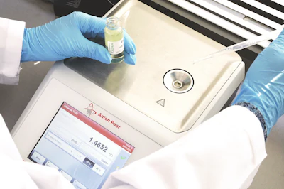 Laboratory technician in blue gloves holding a yellow liquid sample above an Anton Paar analytical instrument during testing