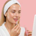 Woman applying pink liquid lipstick while checking mirror with headband on pink background