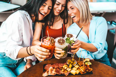 Three women toasting with colorful cocktails and smiling at a social gathering with appetizers on the table