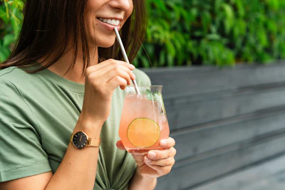 Woman in green shirt smiling while drinking a refreshing citrus beverage with a straw outdoors
