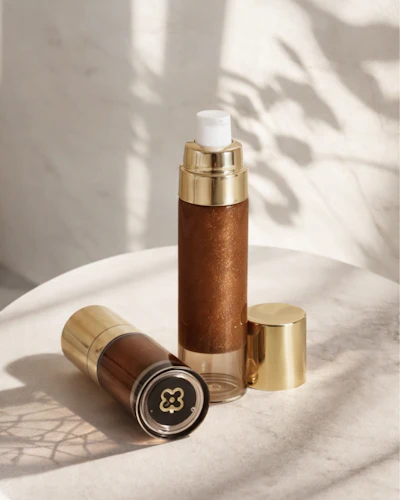 Luxury cosmetic bottles with gold caps and bronze-toned glass containers displayed on white marble with natural sunlight