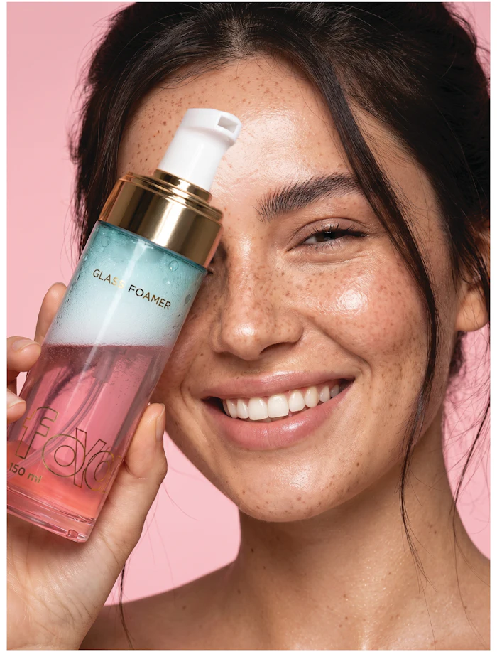 Woman holding GLAS FOAMER facial cleanser bottle, smiling against pink background with visible freckles