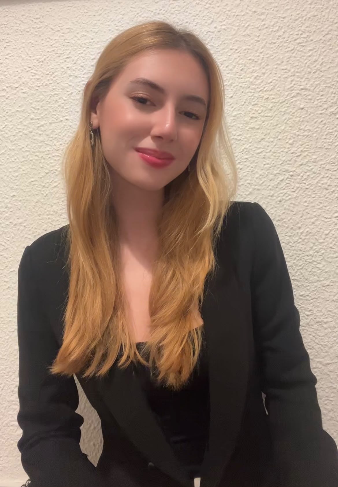 Portrait of a woman with honey-blonde hair wearing a black blazer against a neutral textured wall