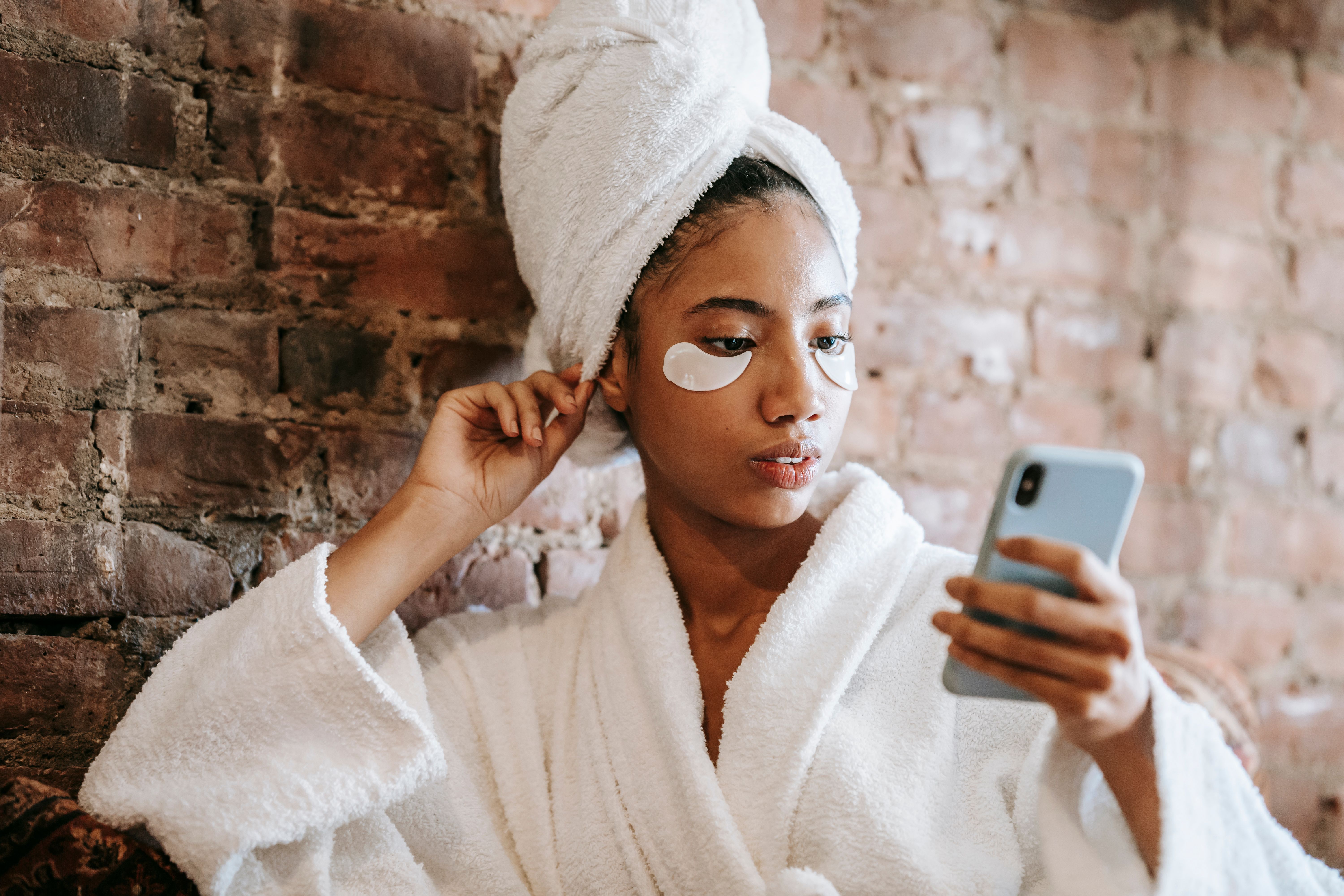 Woman in white bathrobe with under-eye patches checking phone against brick wall