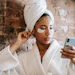Woman in white bathrobe with under-eye patches checking phone against brick wall