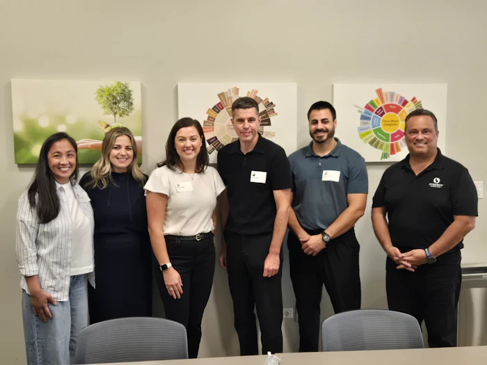 Six professionals smiling at camera in office setting with sustainability graphics on wall behind them