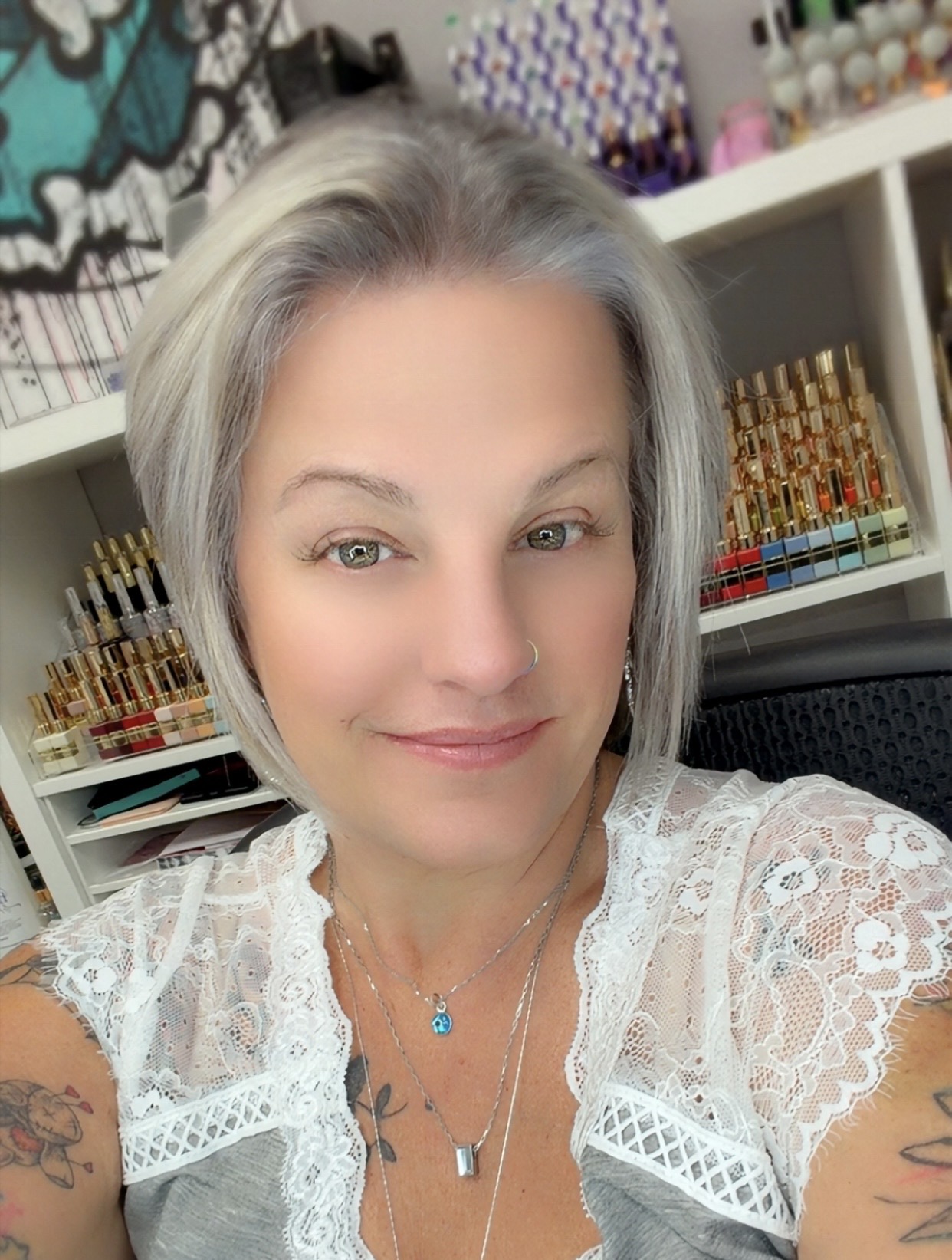 Woman with gray hair wearing white lace top and necklace, smiling in front of organized shelving with art supplies