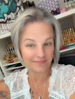 Woman with gray hair wearing white lace top and necklace, smiling in front of organized shelving with art supplies