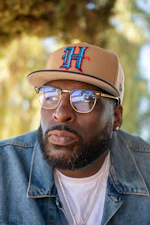 Man wearing tan baseball cap with blue and red H logo, gold-rimmed glasses, and denim jacket outdoors