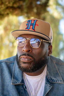 Man wearing tan baseball cap with blue and red H logo, gold-rimmed glasses, and denim jacket outdoors