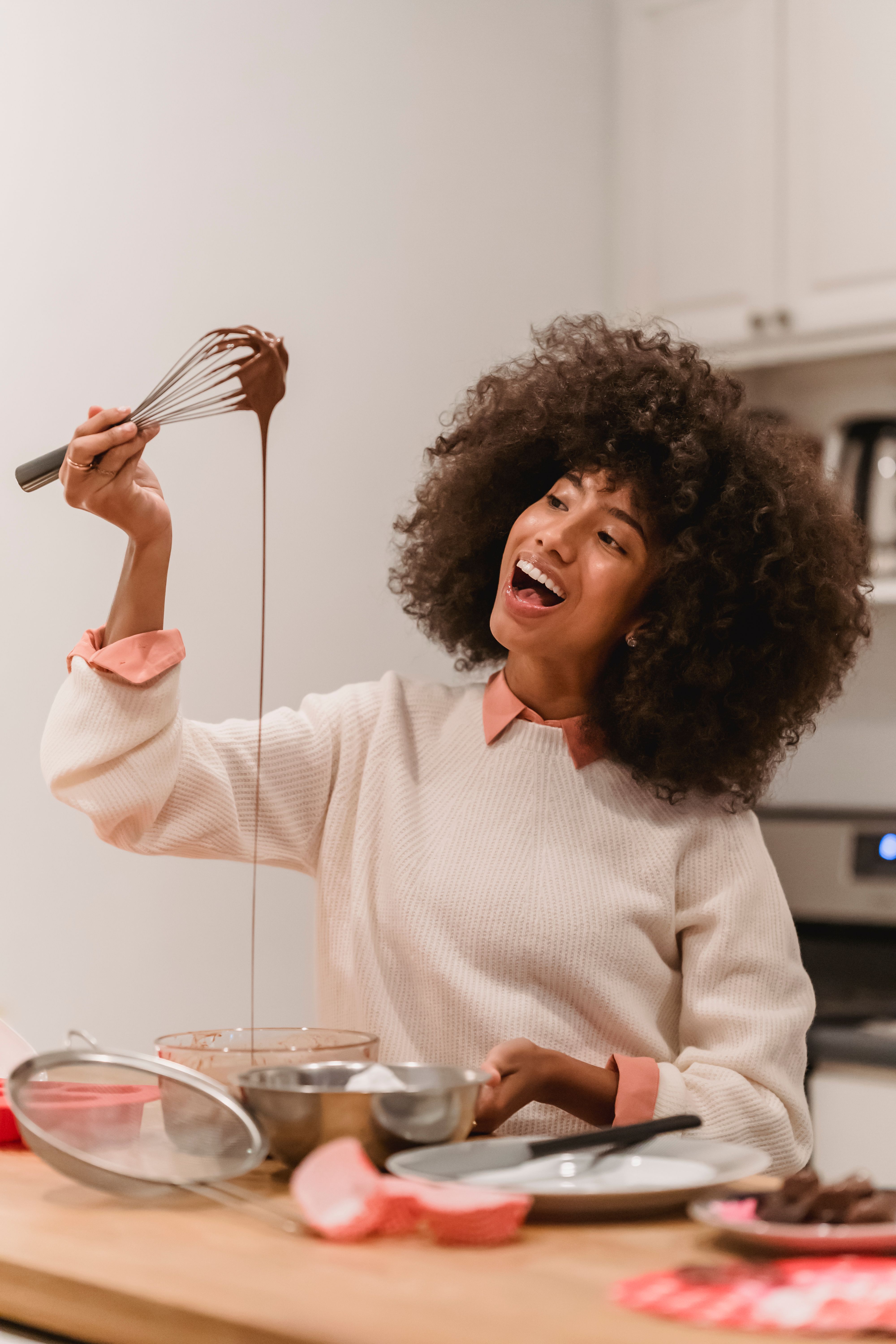 Woman with curly hair laughing while holding a whisk with chocolate batter dripping in a bright kitchen