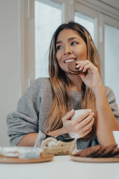 Woman in gray sweater smiling while eating a chocolate cookie at a table by bright windows