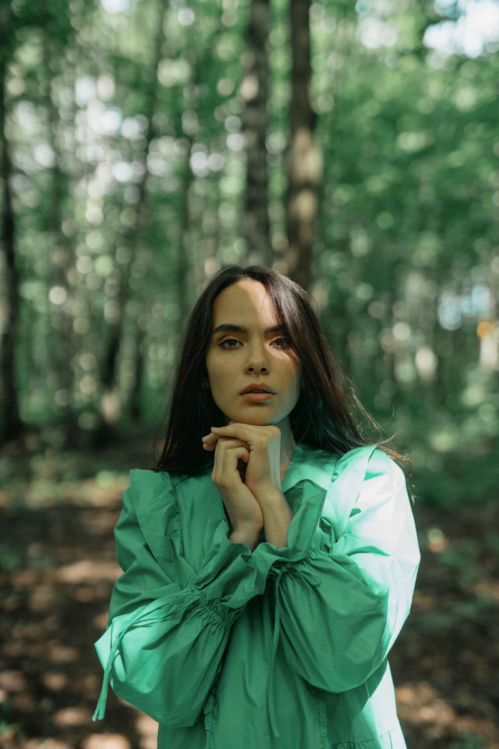 Woman in green jacket standing thoughtfully in a sunlit forest with trees in soft focus behind her