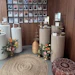 Memorial or tribute display with 32 portrait photographs on wood wall, decorative white ribbed pedestals with floral arrangements, and woven floor cushions.
