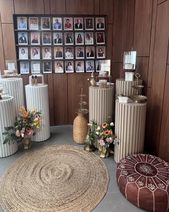 Memorial or tribute display with 32 portrait photographs on wood wall, decorative white ribbed pedestals with floral arrangements, and woven floor cushions.
