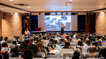 Large audience seated in auditorium facing stage with two presenters and projection screen displaying content during conference or educational event.