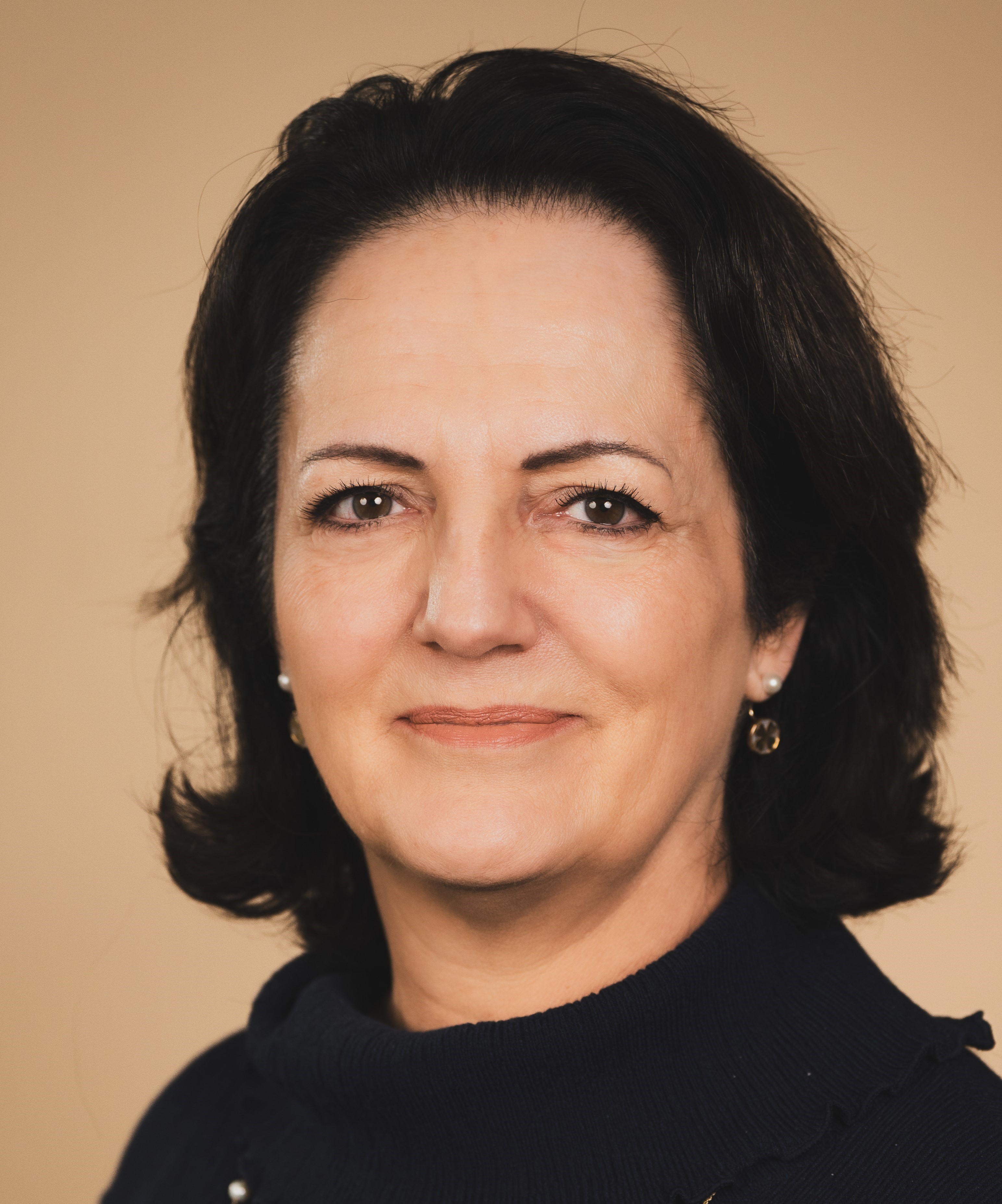 Professional headshot of a woman with dark shoulder-length hair wearing a black top and small hoop earrings against a neutral beige background