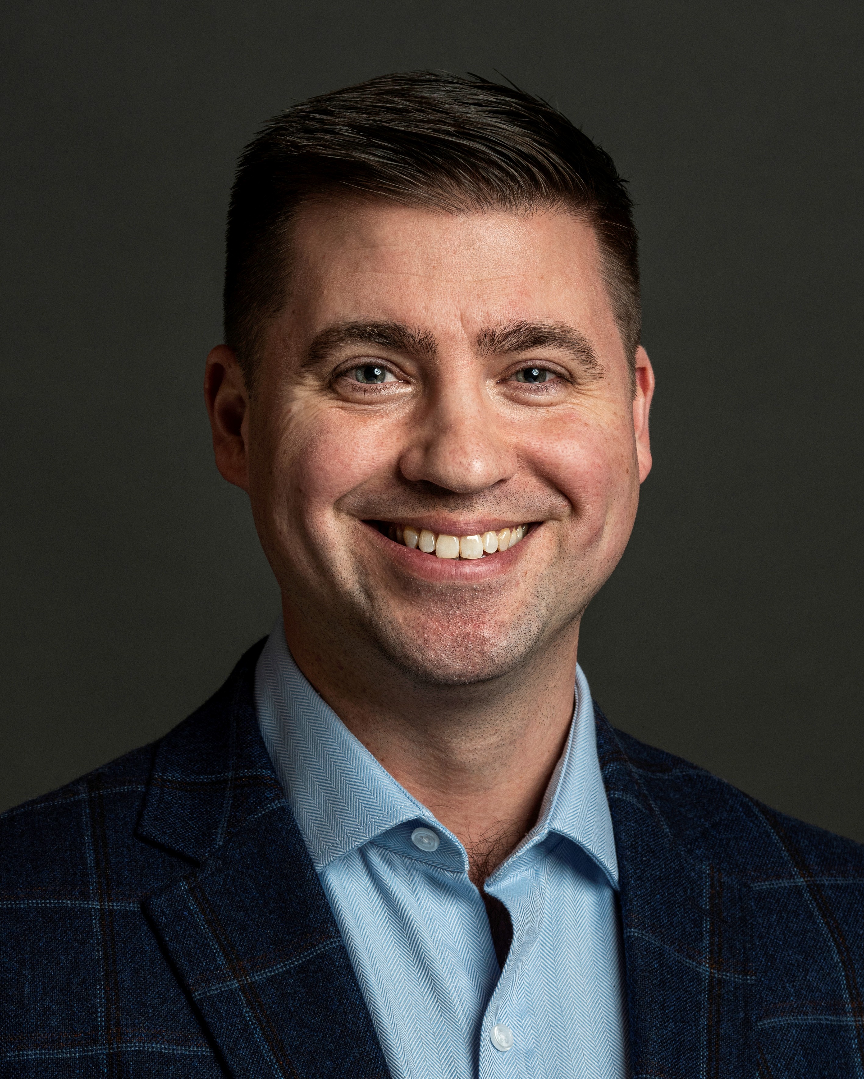 Professional headshot of smiling man in dark blazer and light blue shirt against gray background