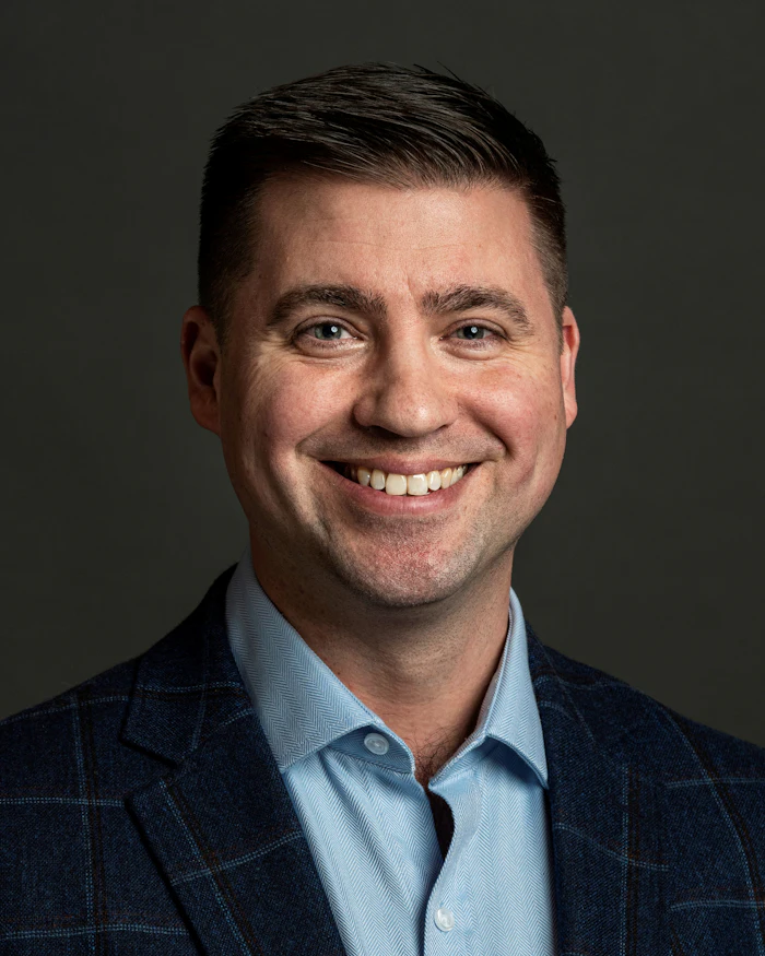 Professional headshot of smiling man in dark blazer and light blue shirt against gray background