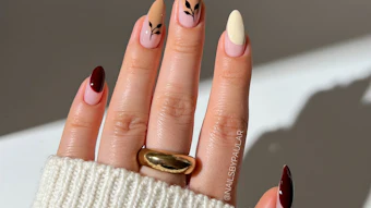 Manicured hand displaying almond-shaped nails with mixed designs including burgundy, nude, cream, and black leaf accents