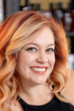 Professional portrait of a smiling woman with wavy reddish-blonde hair wearing a black sleeveless top in an indoor setting