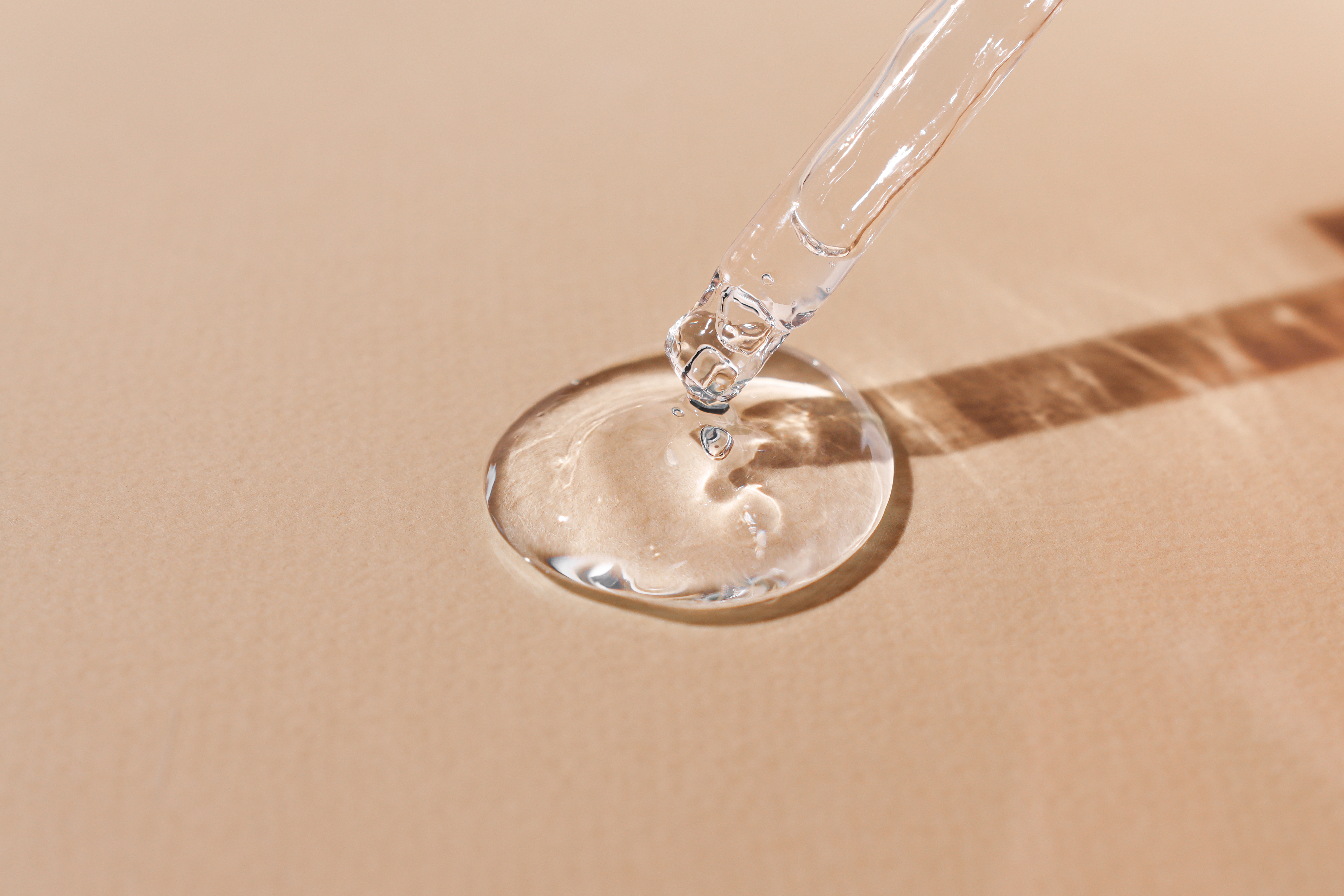 Clear liquid serum dispensed from dropper into glass dish on beige background