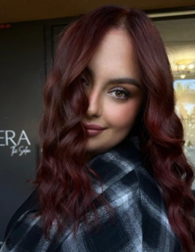 Portrait of a woman with long burgundy wavy hair wearing a dark plaid shirt, looking over her shoulder indoors