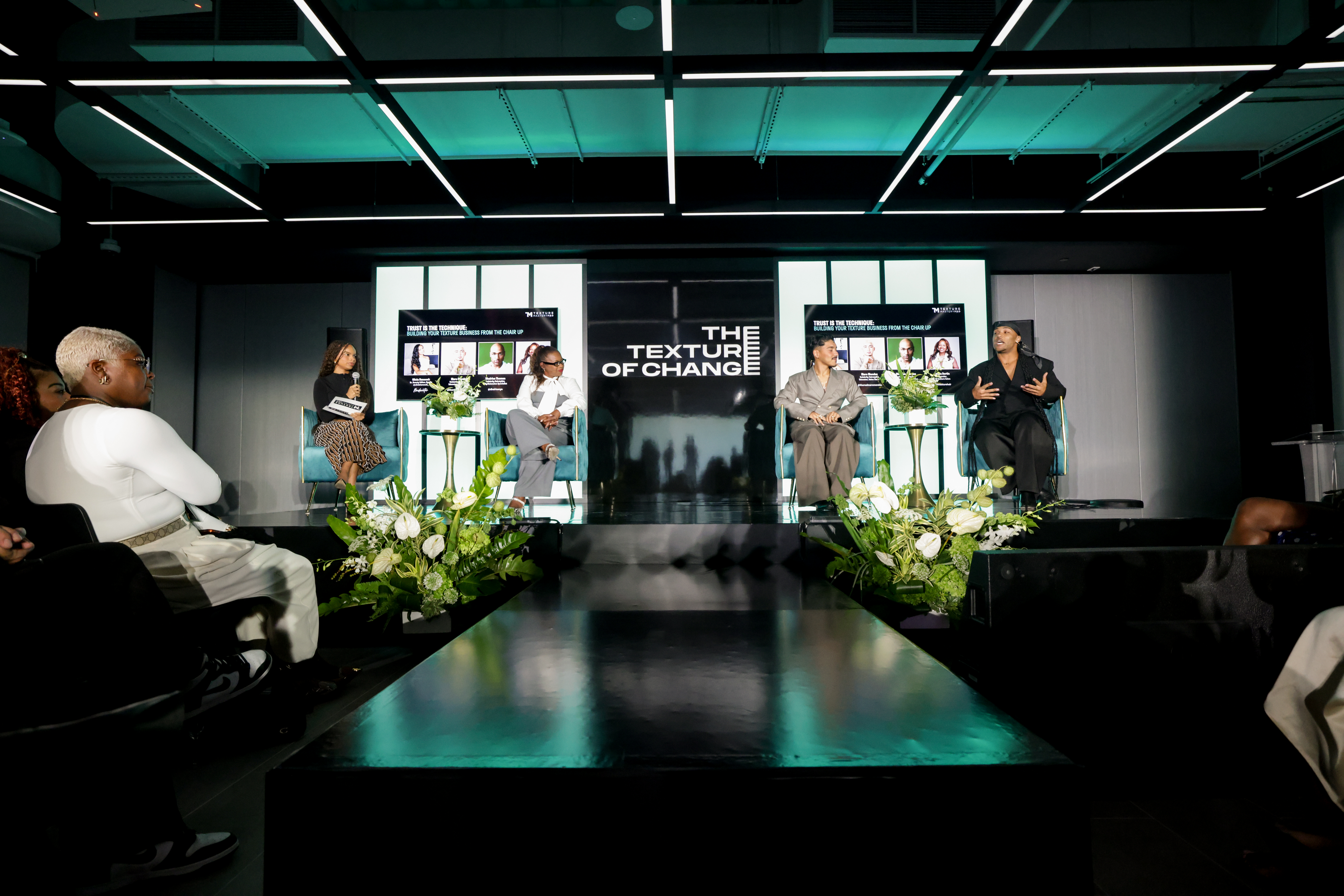 Professional panel discussion event with speakers seated on stage under teal lighting, featuring 'The Texture of Change' backdrop and white floral arrangements.