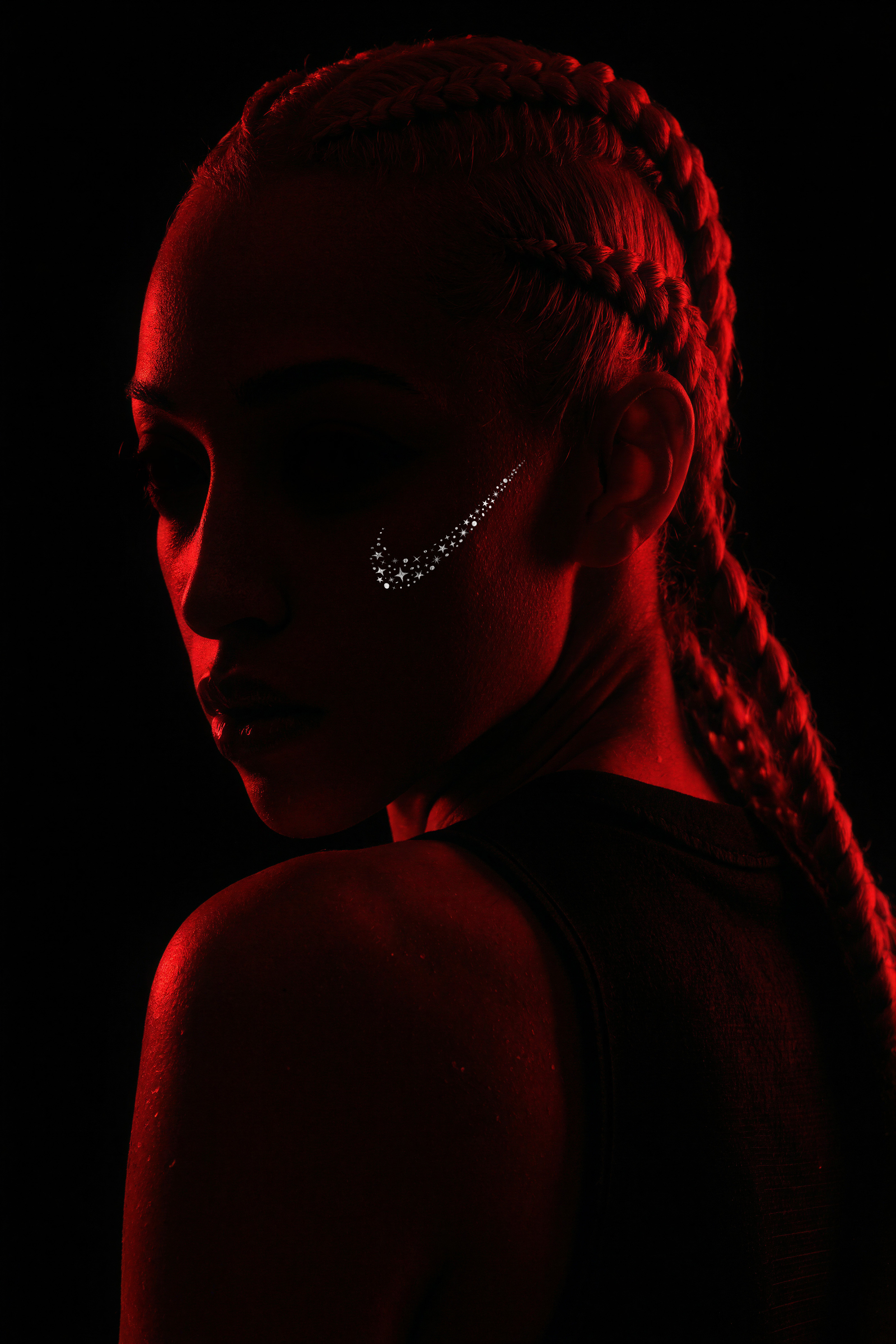 Profile portrait of person with braided hair wearing diamond earring, lit with dramatic red lighting against black background