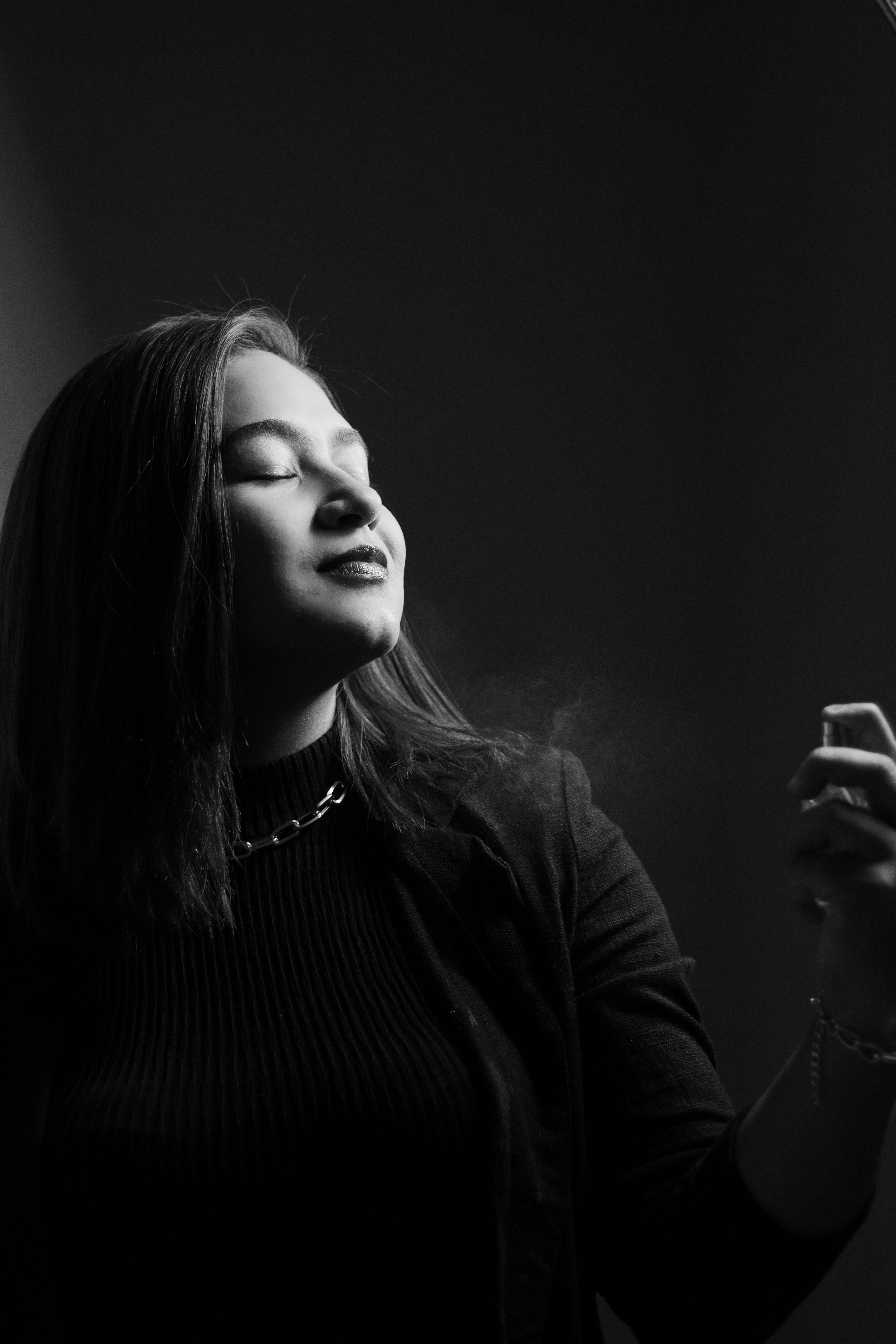 Black and white portrait of a woman with long dark hair, eyes closed, wearing a dark textured jacket and chain necklace against a dark background