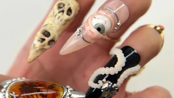 Hand displaying Halloween-themed nail art with skull, eye, and zebra stripe designs in gold, black, and white