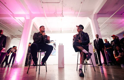 Two men in conversation during a panel discussion at an event with pink ambient lighting and audience members present.